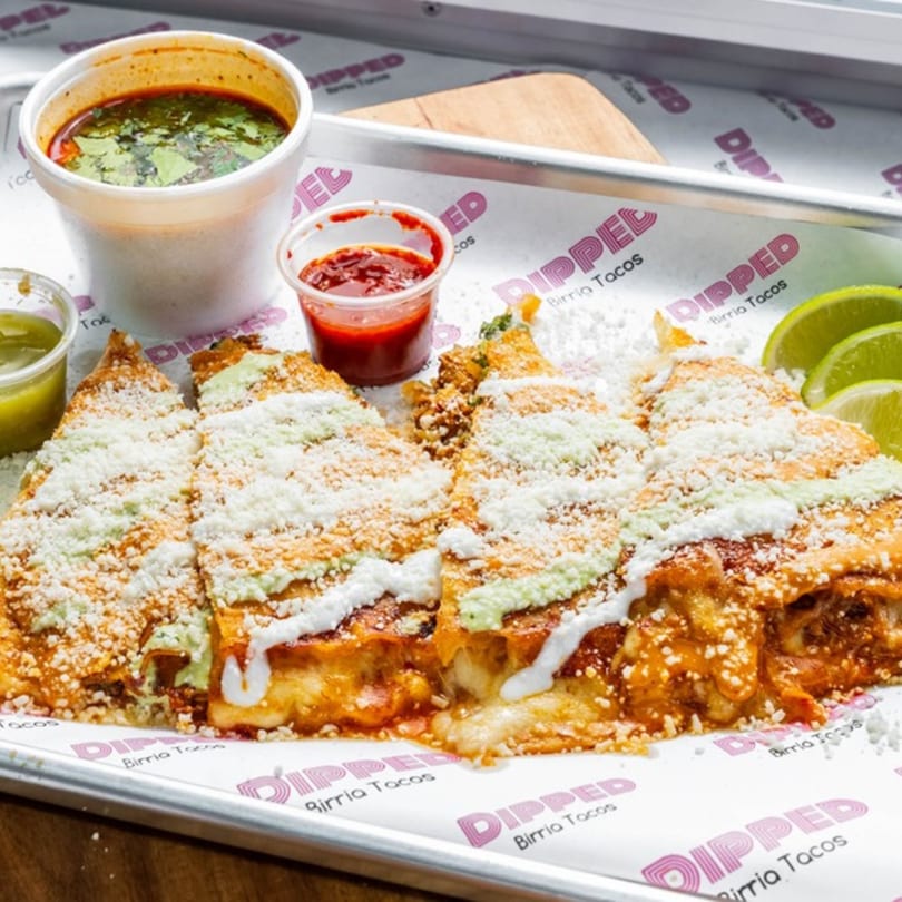 Four Dipped Birria Tacos, garnished with cheese and sauce, served with dipping broth and lime.