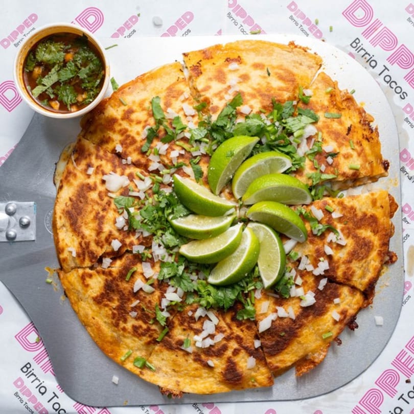 A large, golden-brown birria quesadilla, cut into slices, topped with cilantro, onions, and lime wedges, with a side of consommé.