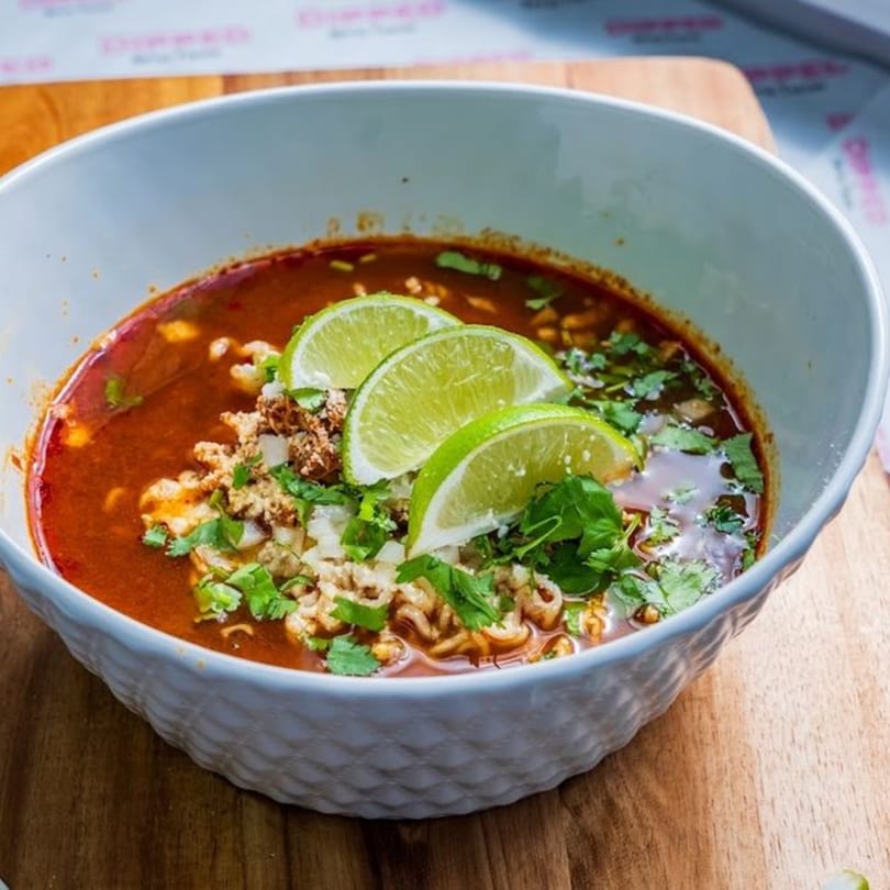 A bowl of Dipped Birria Tacos birria ramen with lime wedges, cilantro, and onions.