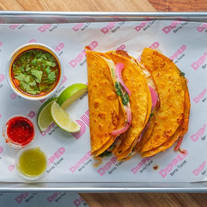 Three Dipped Birria Tacos with consommé, lime wedges, red and green sauces on a tray.
