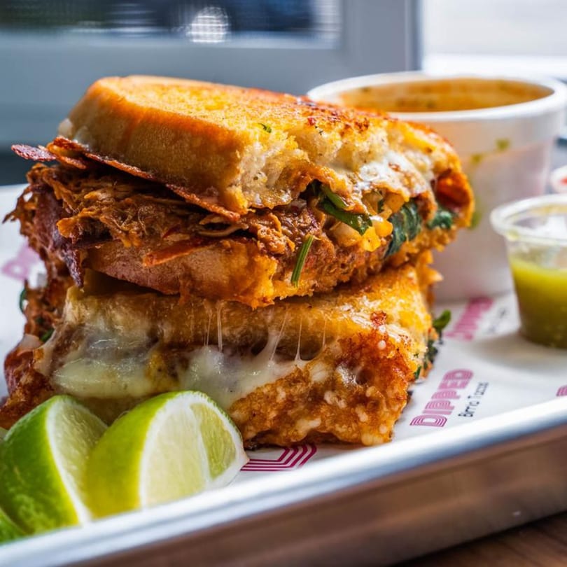 A Dipped Birria Tacos sandwich, with melted cheese and limes, served with dipping sauce.