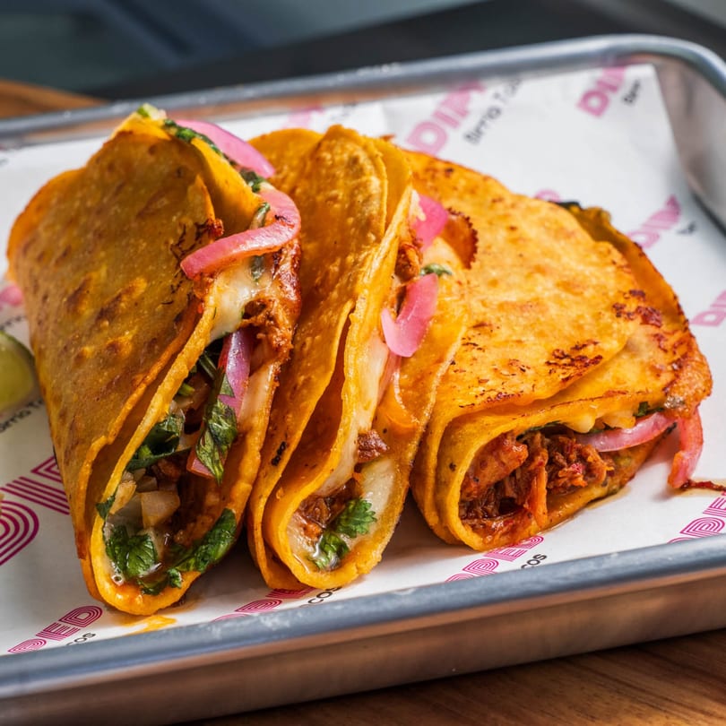 Three Dipped Birria Tacos, filled with meat, cheese, cilantro, and pickled red onions, on a metal tray.
